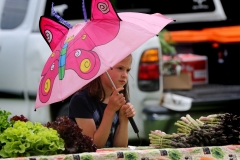 Bethany Roberts, 7, protects herself from occasional drizzle at her family's produce stand during Busy Kids Day at the Mukwonago Farmer's Market at Field Park on June 6 that offered a wide variety of interactive activities for children.